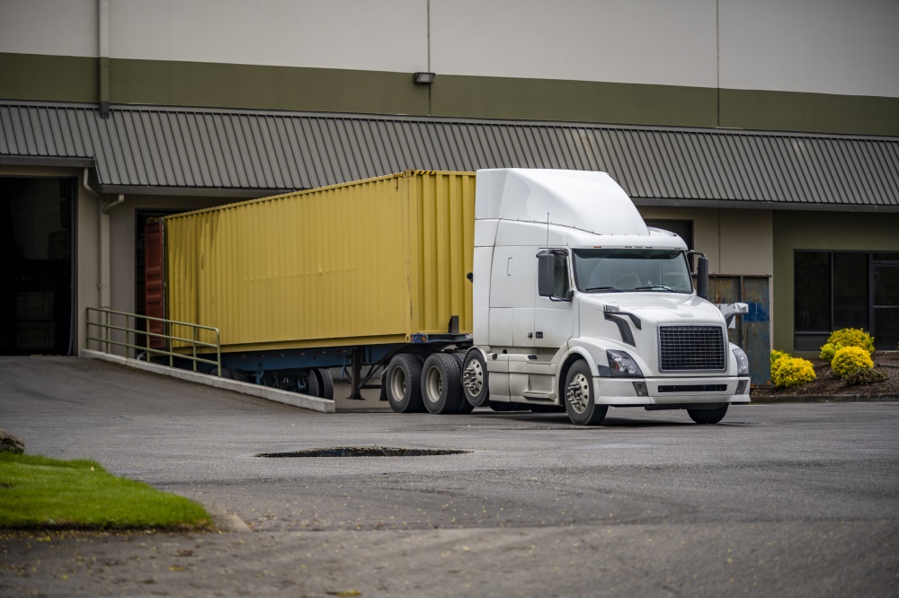 White-big-rig-professional-semi-truck-with-container-trailer-unloading-cargo-standing-in-wareho.jpeg